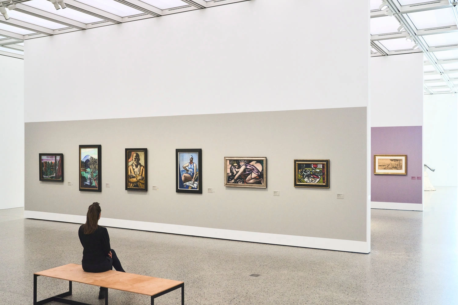 Person sitting on a bench in a modern museum hall, looking at a series of framed paintings on the wall.
