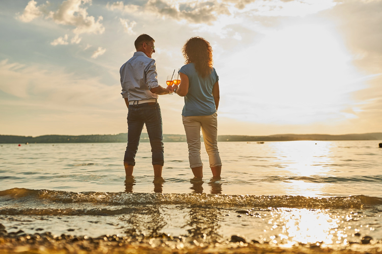 Ein Mann und eine Frau stehen knöcheltief im Wasser eines Sees und stoßen mit einem Glas an.