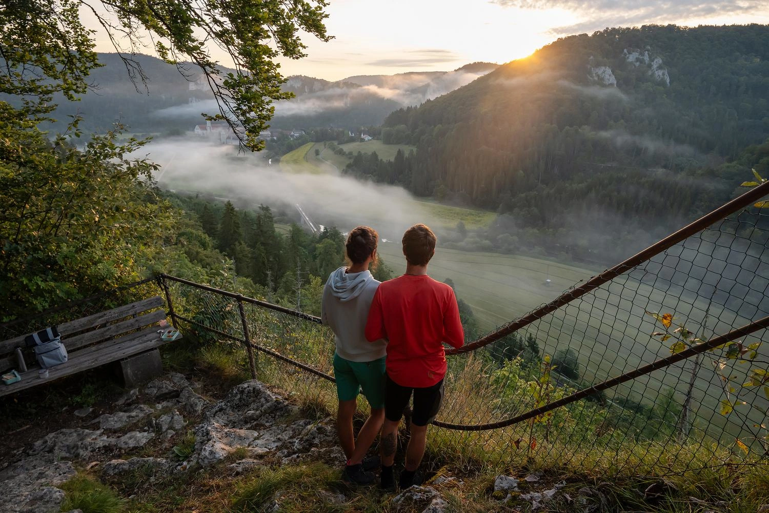 Zwei Wanderer stehen an einem Aussichtspunkt und blicken in ein Tal, durch das Nebelschwaden wabern. Die Sonne geht gerade auf.