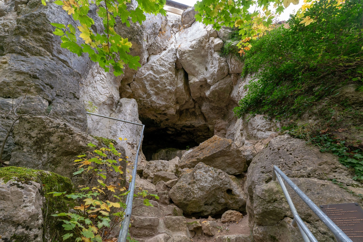 Stone cave entrance with large boulders, surrounded by green vegetation and a railing along the ascent.