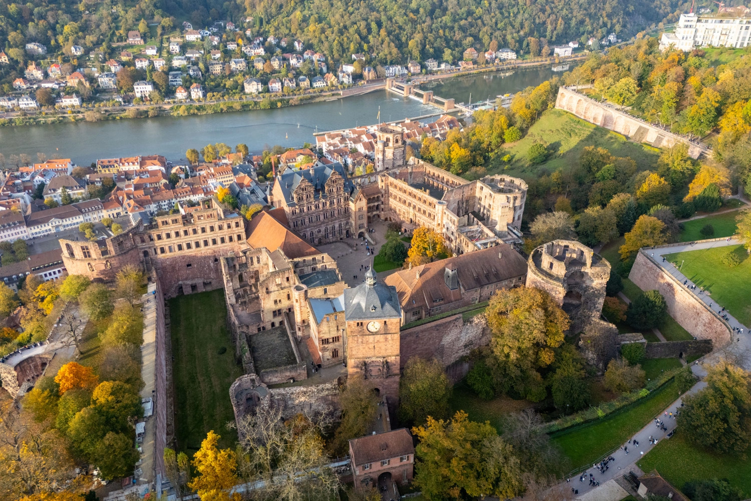 Aerial view of Heidelberg Castle in autumn, surrounded by colorful foliage, with a view of the old town, the Neckar Bridge, and the river; wooded hills and residential buildings in the background.