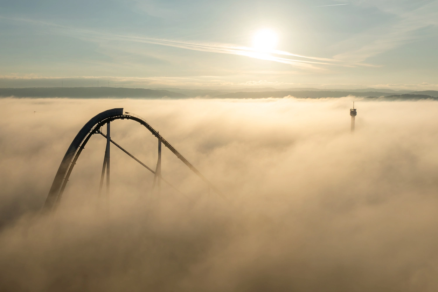 A roller coaster and a tower rise out of thick fog at sunrise.
