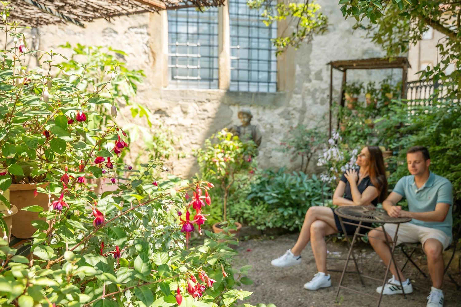 Zwei Personen sitzen an einem kleinen Tisch in einem sonnigen Garten mit üppigen grünen Pflanzen und pinken Blüten vor einer alten Steinmauer.