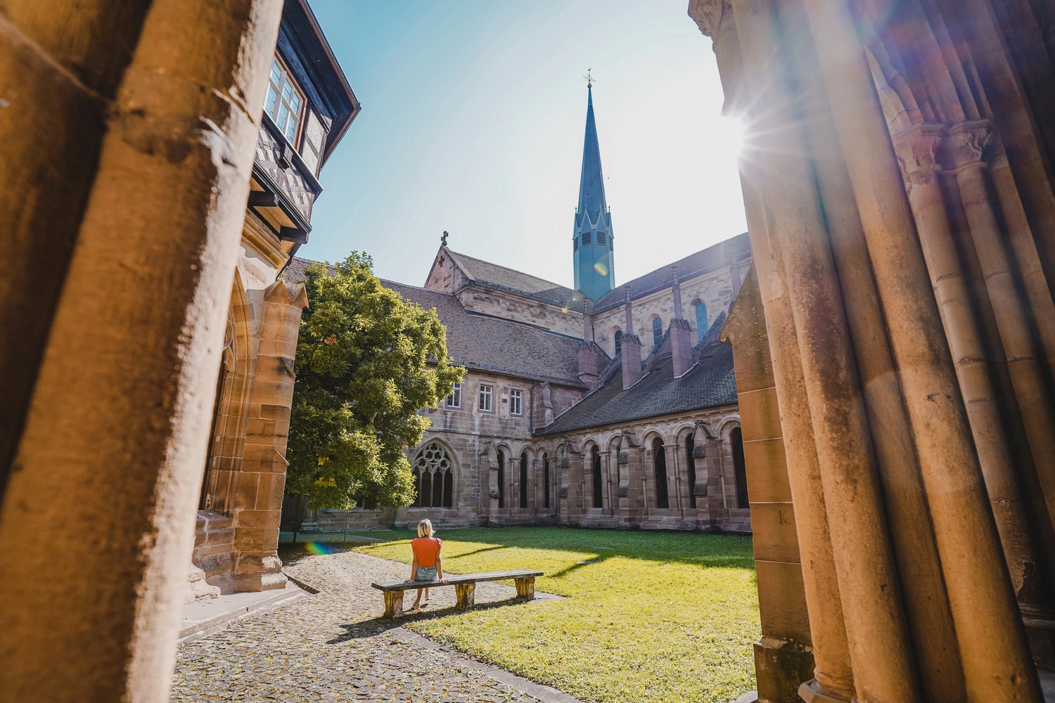 Sonnendurchfluteter Klosterhof mit Arkaden; eine Person sitzt auf einer Bank im Grünen vor den historischen Gebäuden und dem Kirchturm.