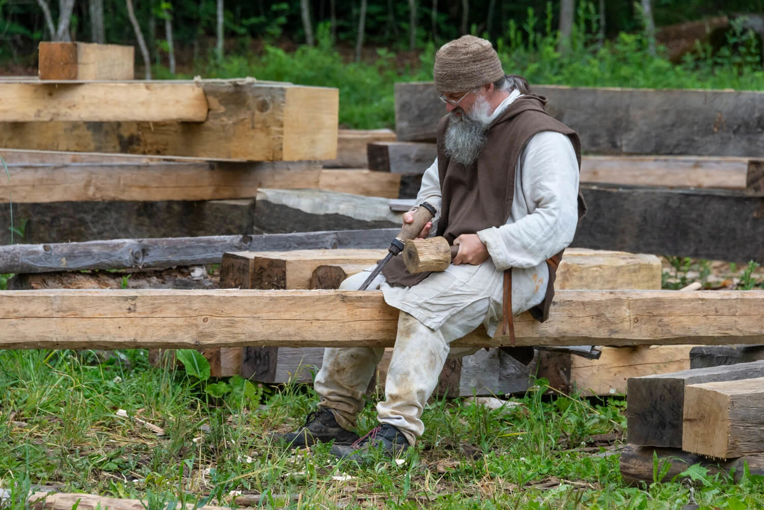 A man in medieval clothing sits on a square sawed tree trunk. There are more tree trunks in the background. The man is holding a file.