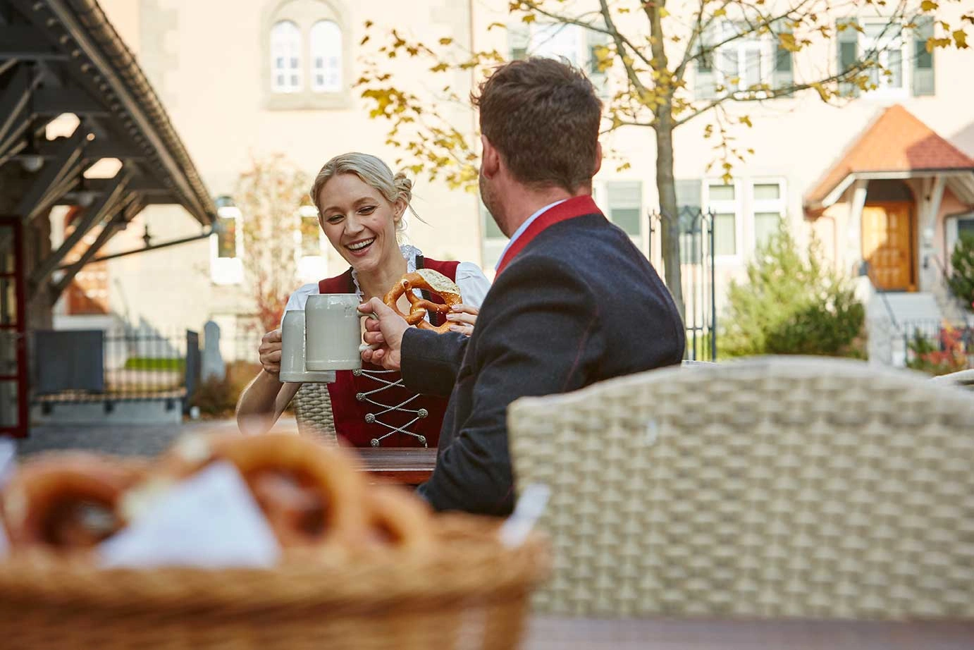 Zwei Personen in Tracht sitzen in einem Biergarten und stoßen mit Bierkrügen an.