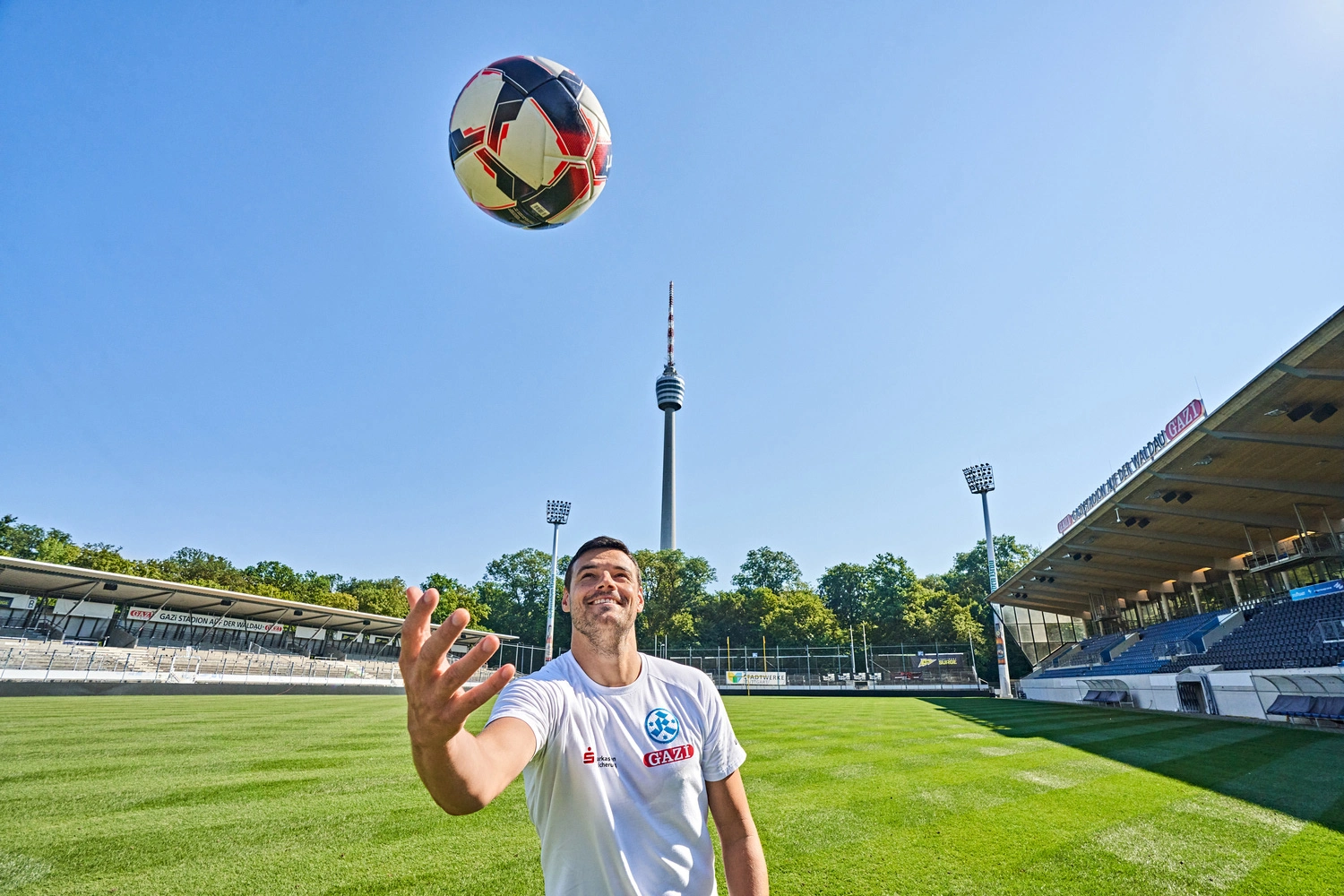 Soccer player kicks a ball into the air on the lawn of the Kickers Stadium in Stuttgart, with the TV tower in the background under a clear blue sky.