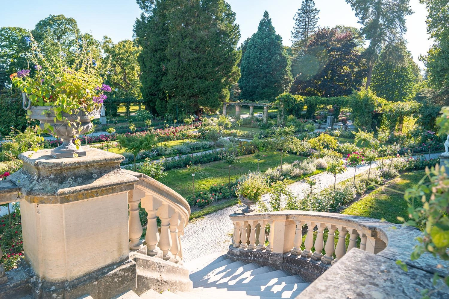 Blick von einer steinernen Freitreppe auf einen gepflegten, blühenden Garten mit Blumenbeeten, Wegen und hohen Bäumen im Sonnenlicht.