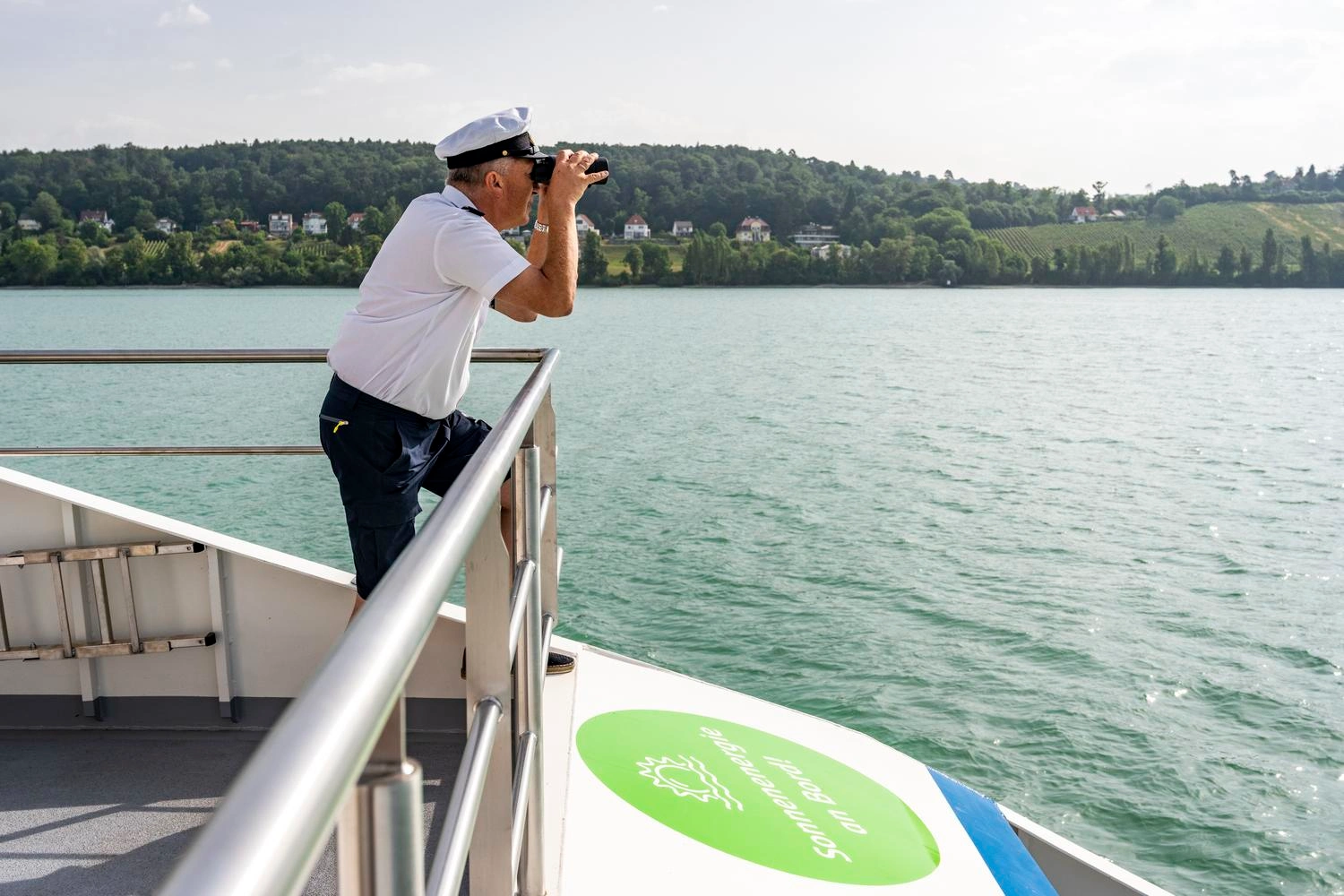 A captain in uniform stands on his ship and looks out over the water through binoculars.