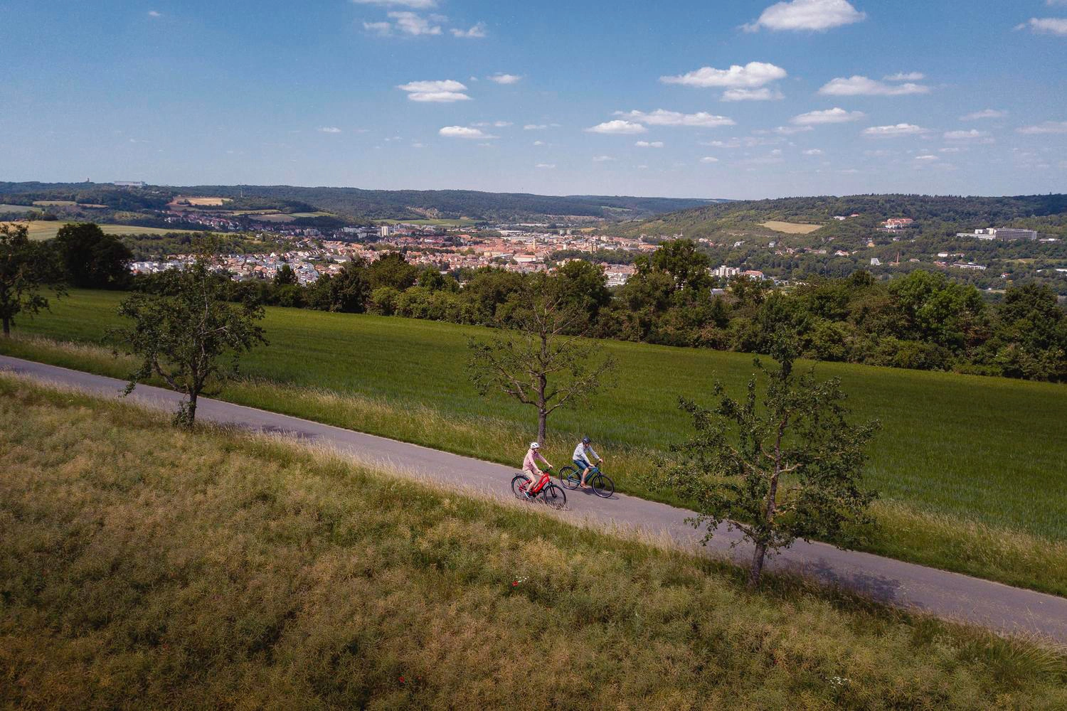 Zwei Radfahrer fahren auf einem Radweg. Im Hintergrund befindet sich in einer Talmulde eine Stadt.
