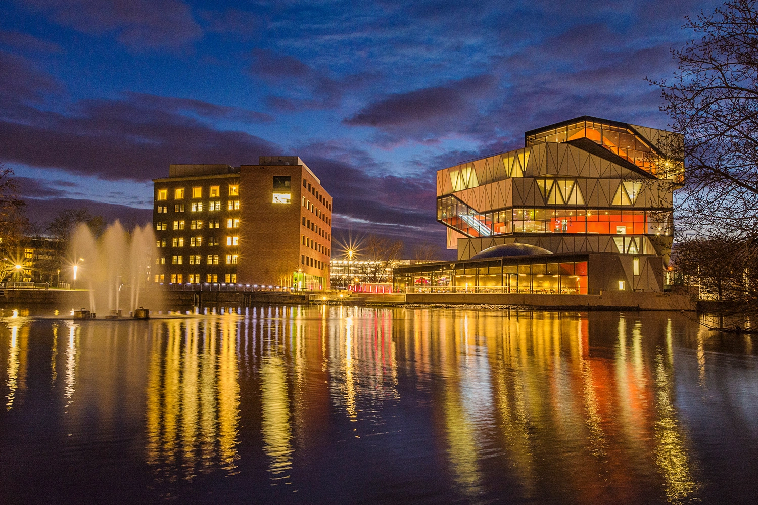 Moderne, geometrisch gestaltete Gebäude der experimenta Heilbronn am Wasser, beleuchtet bei Nacht; bunte Lichtreflexionen spiegeln sich im ruhigen See, links sprudeln Wasserfontänen.