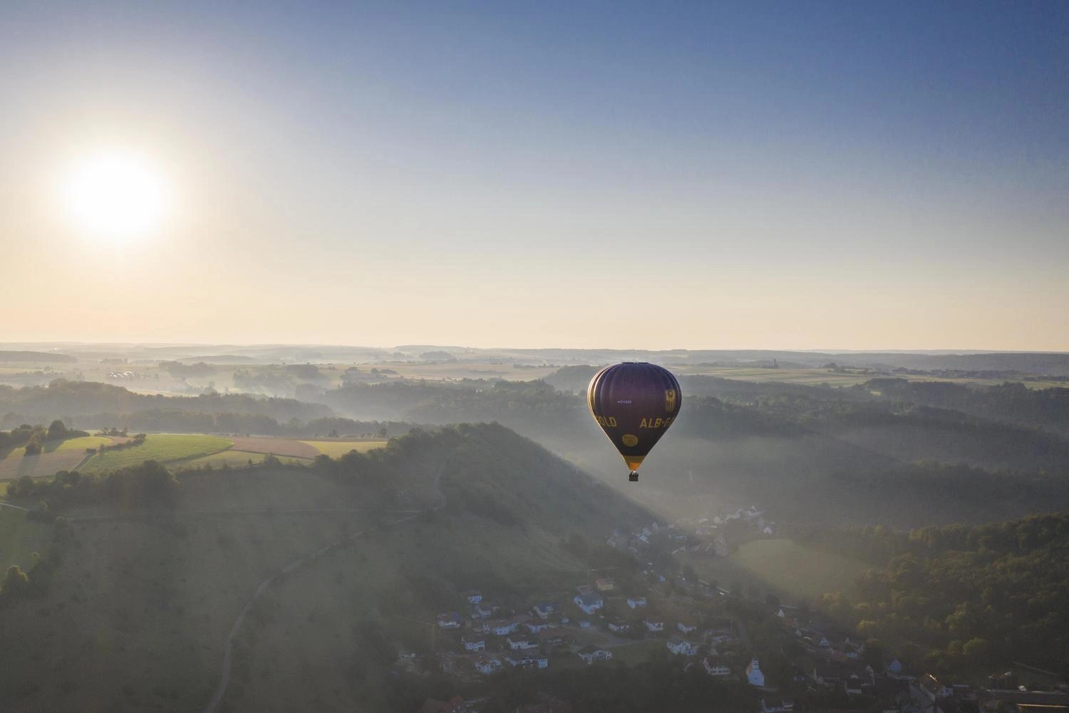 A hot air balloon floats across a hilly landscape toward the sun.