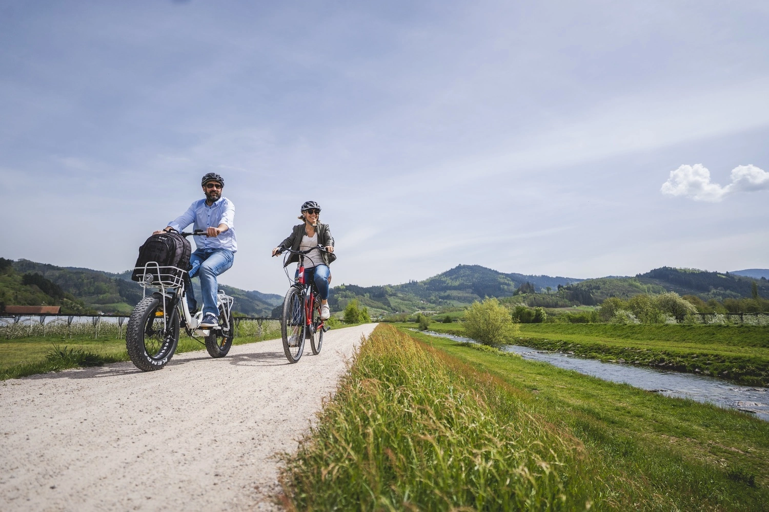 Zwei Radfahrer fahren auf einem erhöhten Radweg neben einem Fluss. Eines der Räder ist ein Fatbike.