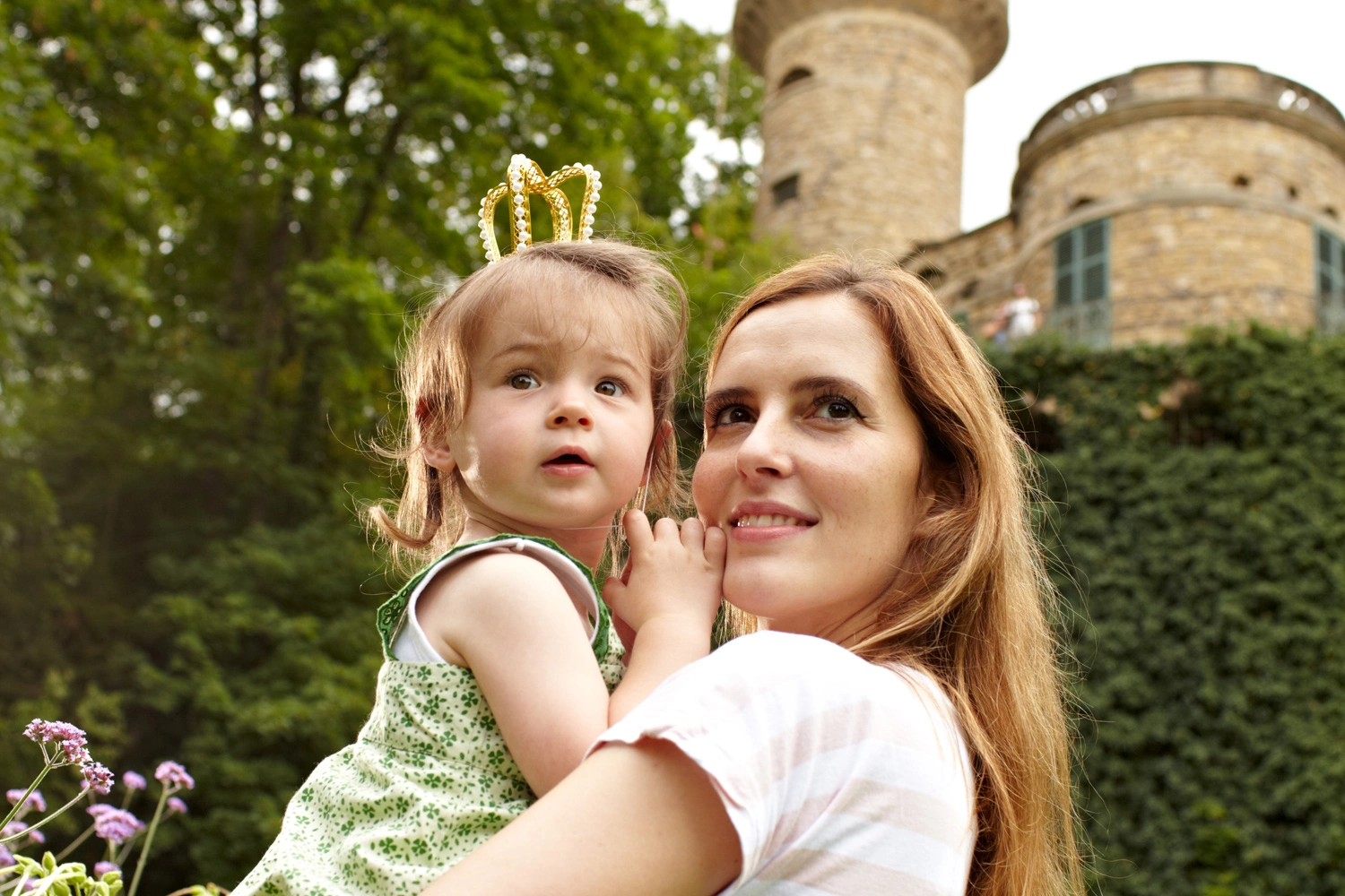 A child wearing a golden crown is carried by a person, with a historic castle and green vegetation in the background.
