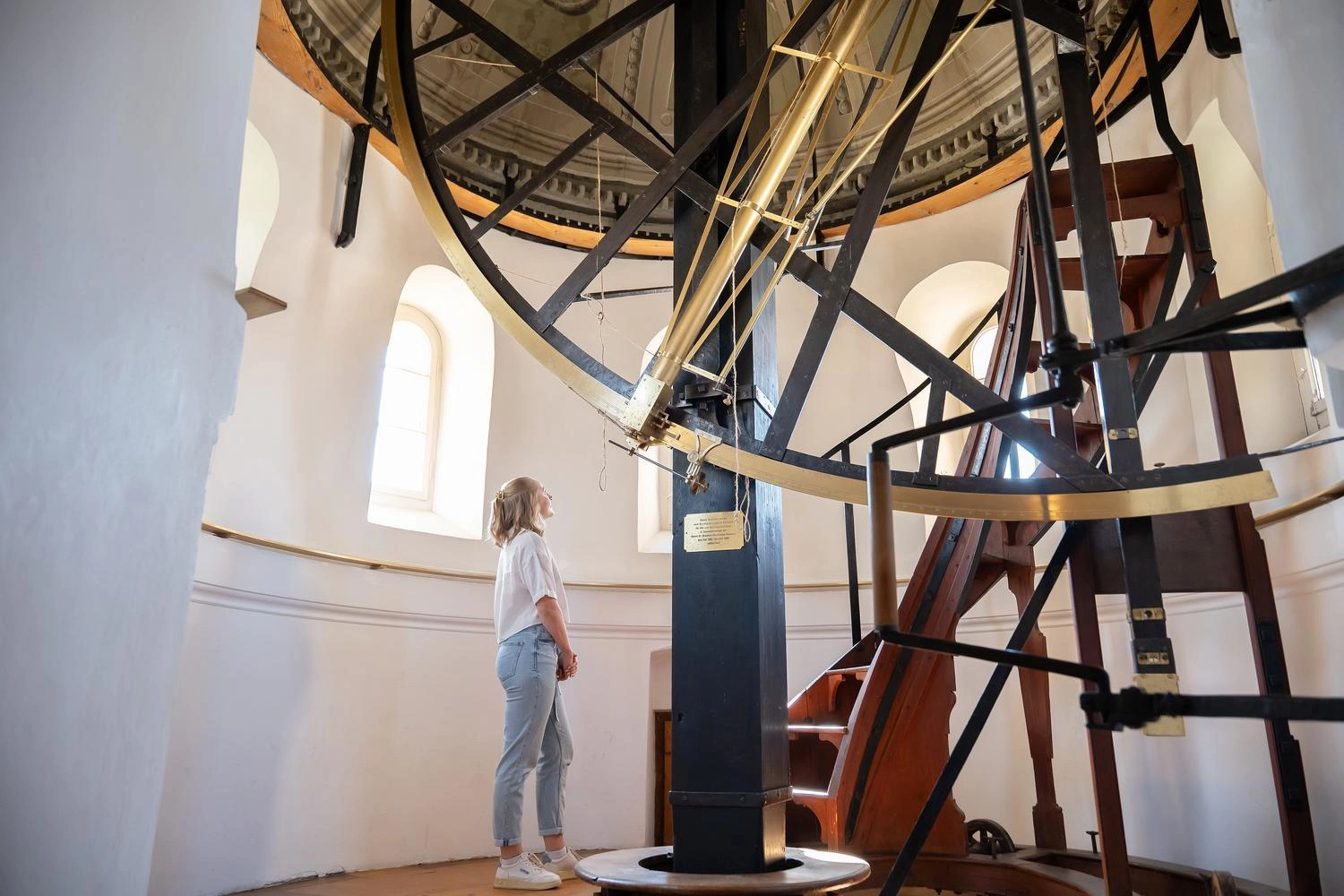 Person betrachtet ein großes historisches Teleskop in einer Sternwarte mit weißen Wänden und Holztreppe.