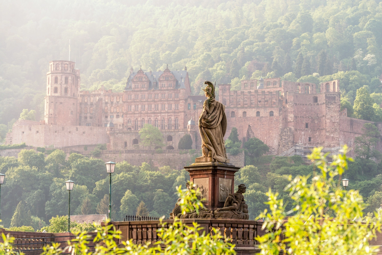 View of Heidelberg Castle surrounded by green hills, with the Minerva Monument on the Old Bridge in the foreground.