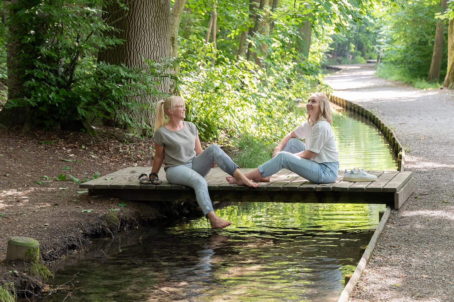 Zwei Personen sitzen auf einem Holzsteg über einem Bach in einem grünen Wald und tauchen ihre Füße ins Wasser.