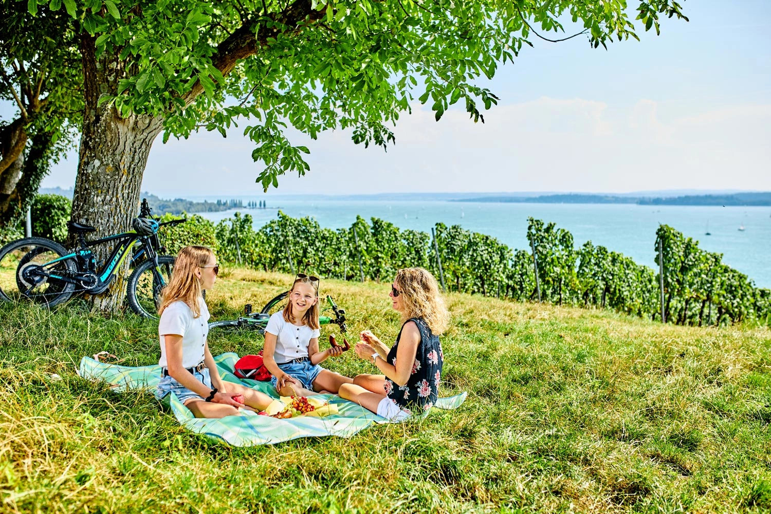 Eine Frau sitzt mit zwei Kindern auf einer Picknickdecke in den Weinbergen.