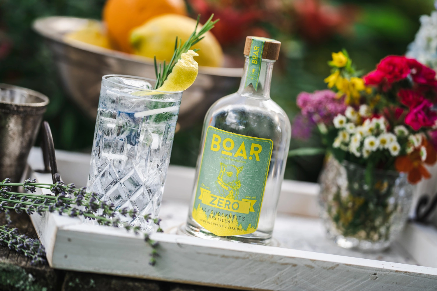 The non-alcoholic distillate BOAR ZERO stands on a table together with flowers, fruit and tonic water in a glass.