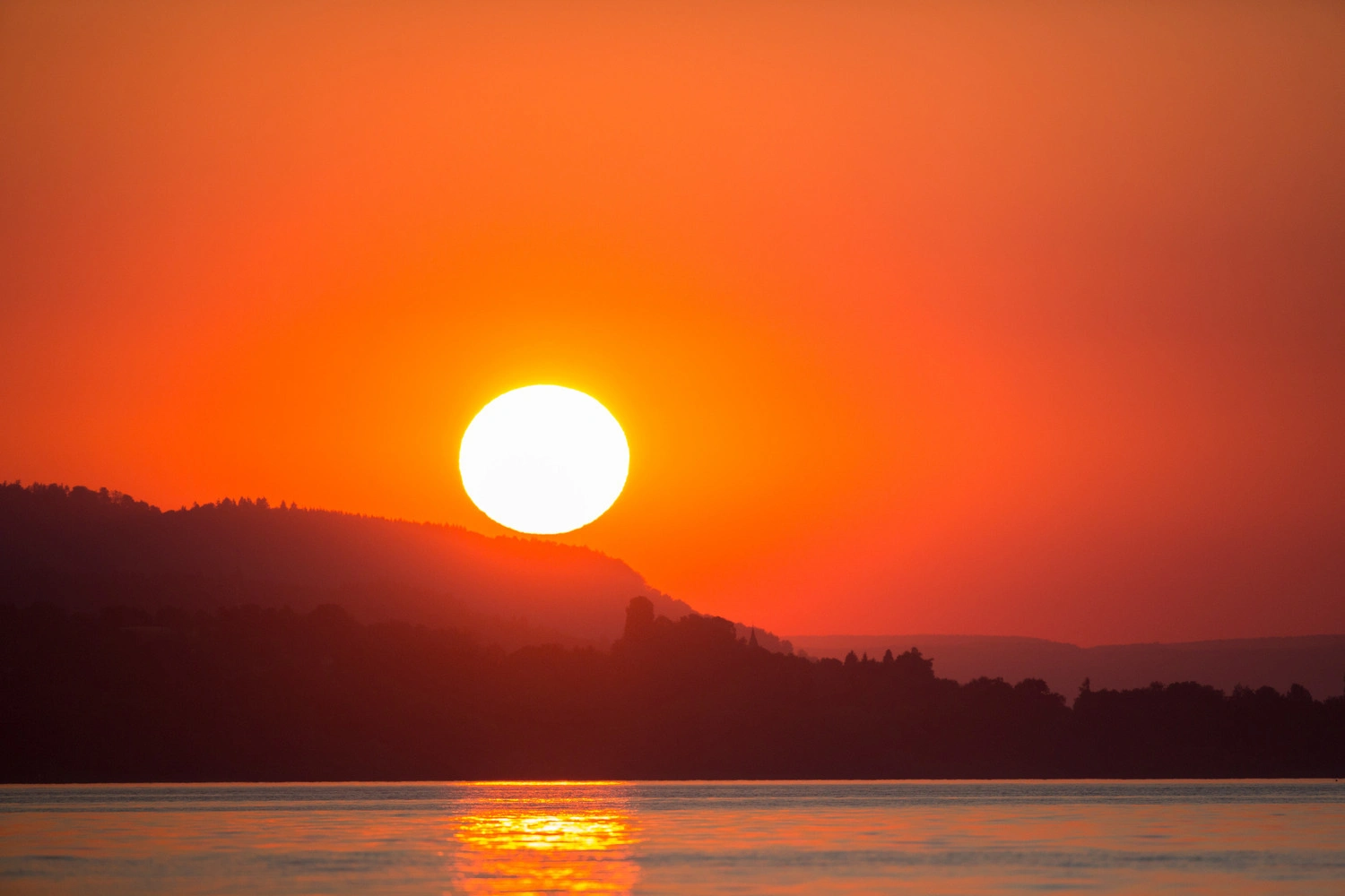 Sonnenuntergang über einem See mit spiegelnder Wasseroberfläche, die Sonne steht knapp über den dunklen Hügeln am Horizont, der Himmel leuchtet in warmen Orange- und Rottönen.