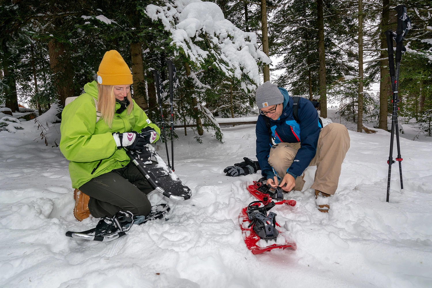 Two people kneel in the snow and put on their snowshoes.