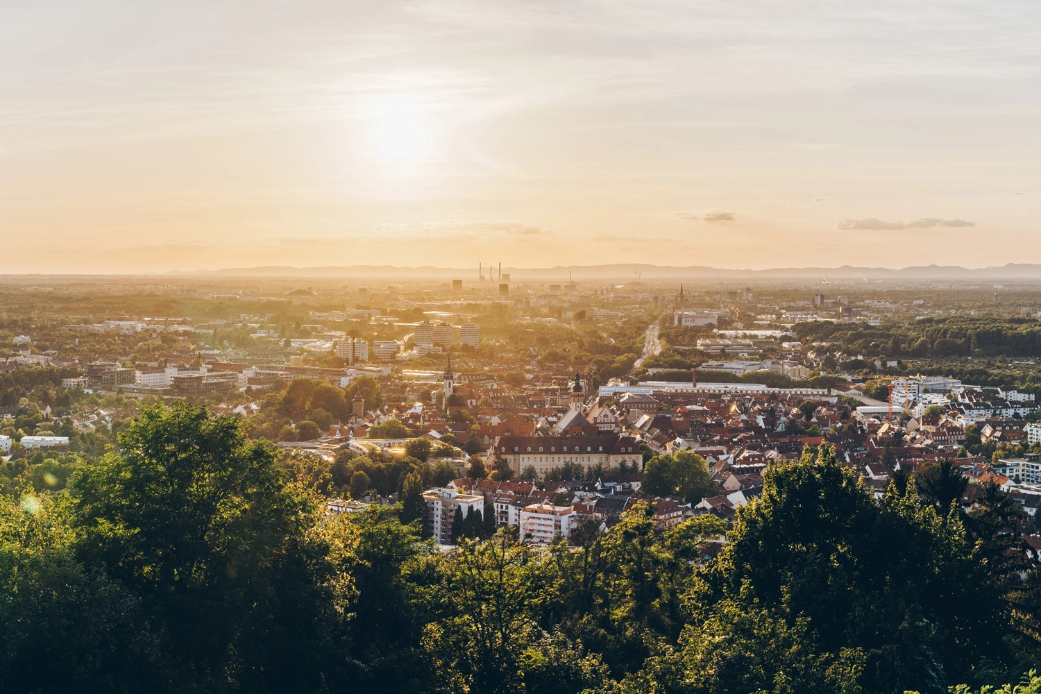 Sunset over Karlsruhe: A view from the wooded Turmberg of the city with its red roofs, churches, and expansive plains bathed in the warm evening light.