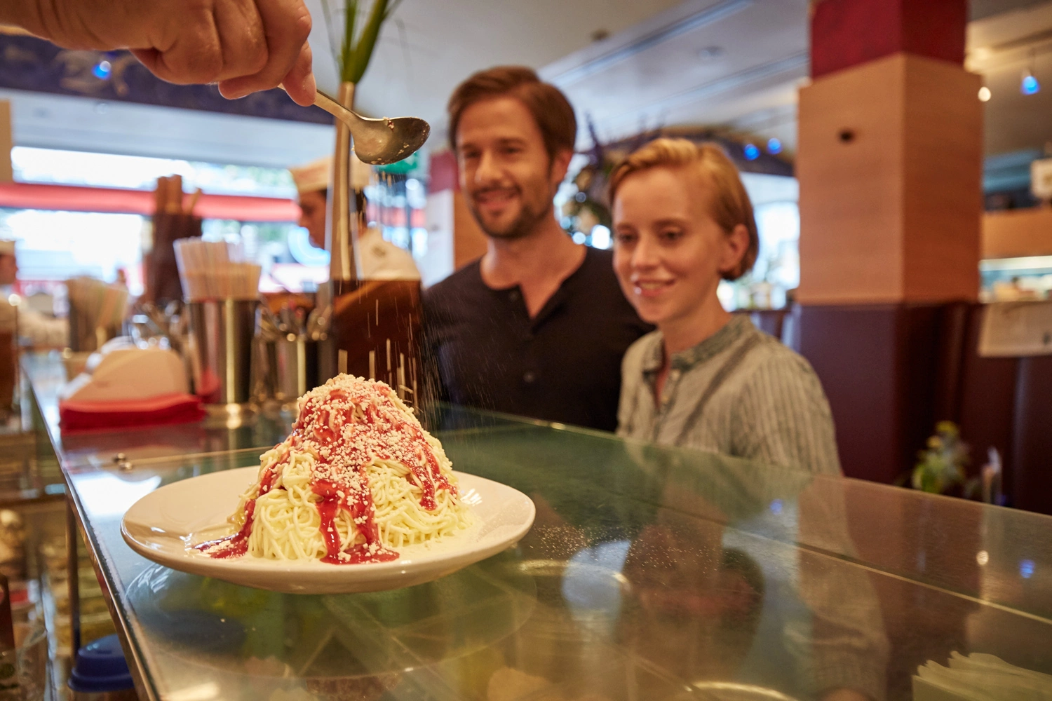 Spaghetti-Eis auf weißem Teller mit Erdbeersoße und geraspelter weißer Schokolade, während eine Hand Kakaopulver darüber streut – Szene in einer Eisdiele am Tresen.