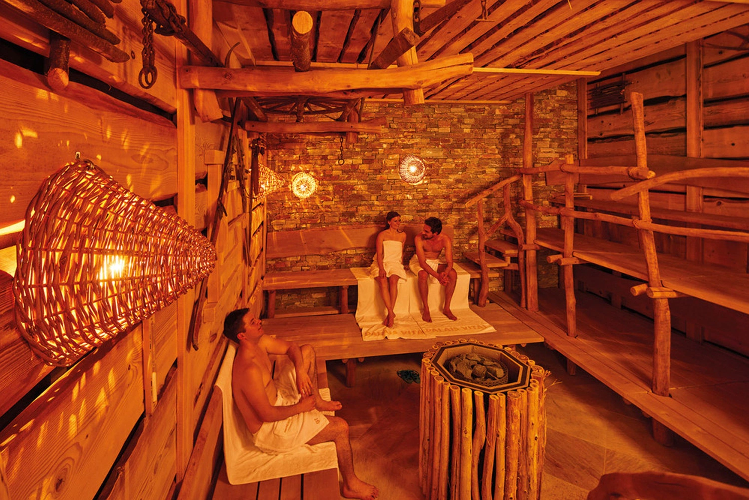 In einer großen Sauna sitzen drei Personen auf Handtüchern auf Holzbänken und ruhen sich aus.
