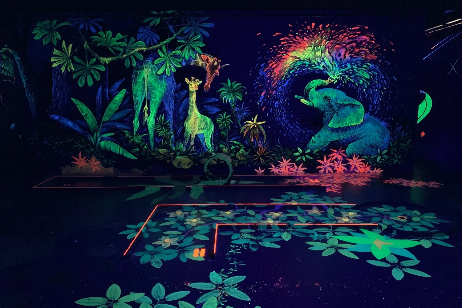 Schwarzlicht-Minigolfbahn mit leuchtenden Dschungelmotiven, Giraffe und Elefant in Neonfarben.