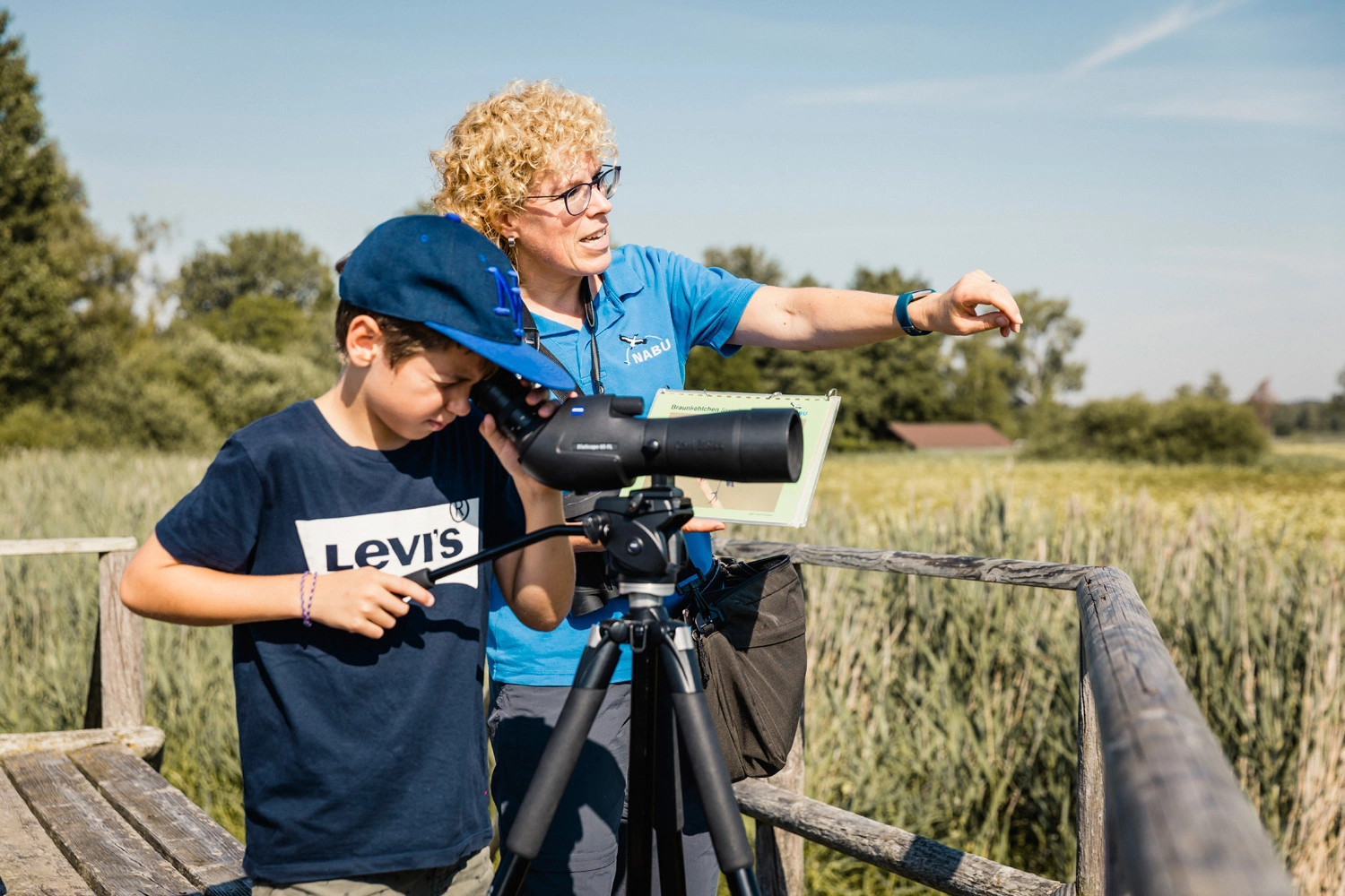 Eine Frau und ein Kind stehen vor einem Fernglas. Der Junge schaut durch das Fernglas und die Frau zeigt mit ihrem Finger auf etwas in der Landschaft.