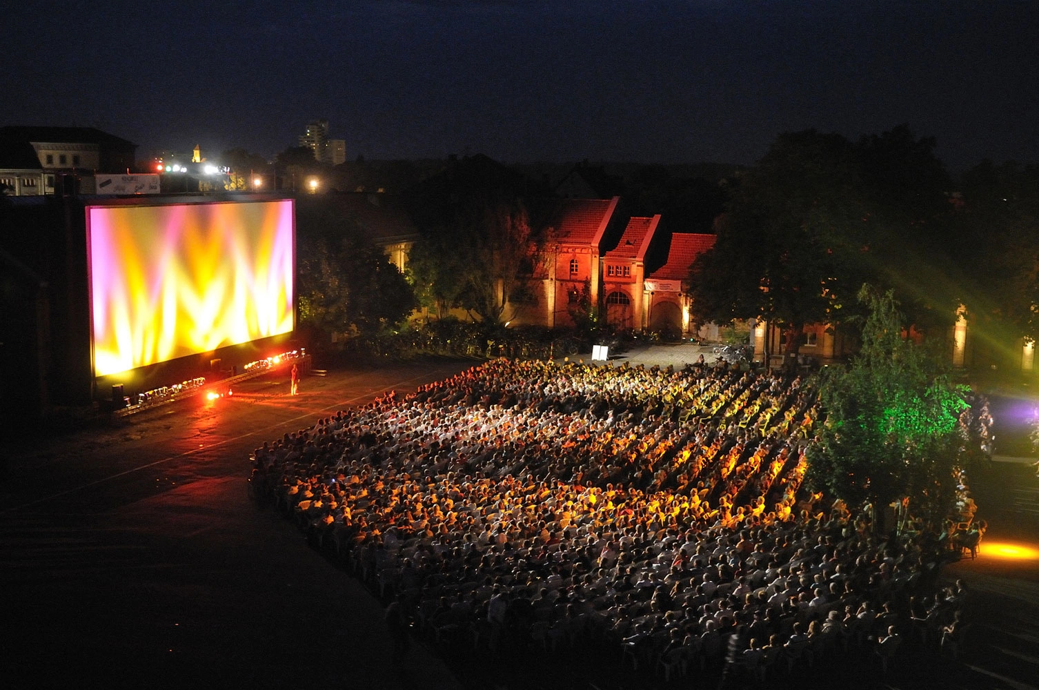 A large crowd gathered for an open-air movie screening the night before, in front of an illuminated screen and historic buildings.