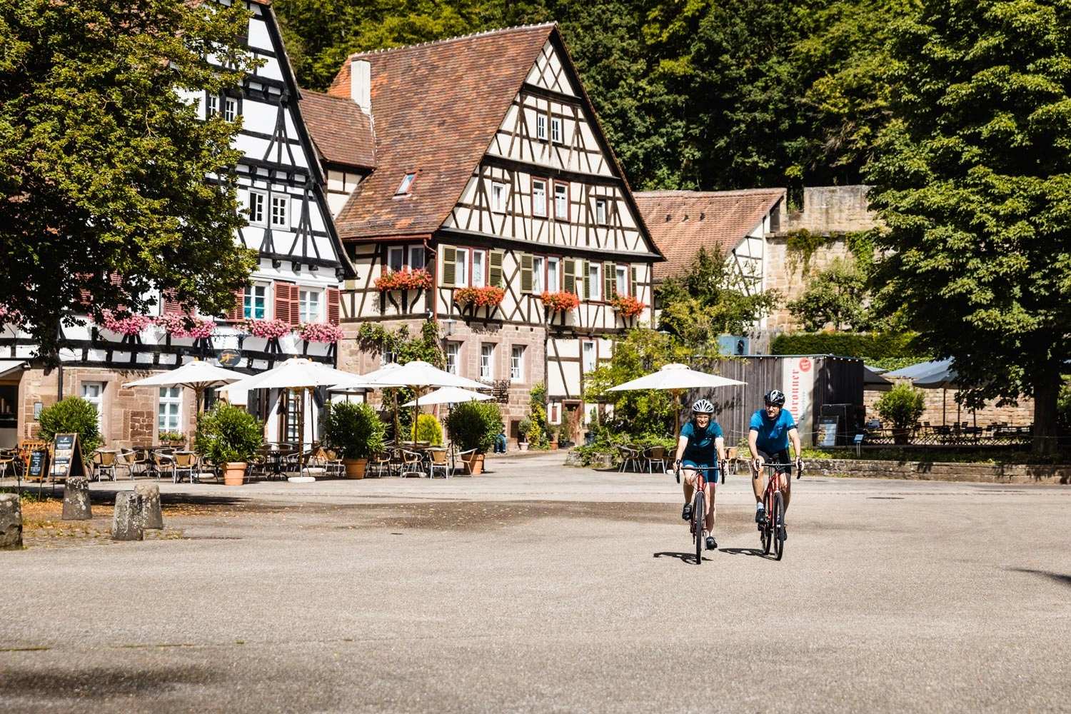 Ein Radfahrer und eine Radfahrerin radeln durch einen idyllischen Ort mit Fachwerkhäusern und großen Bäumen.