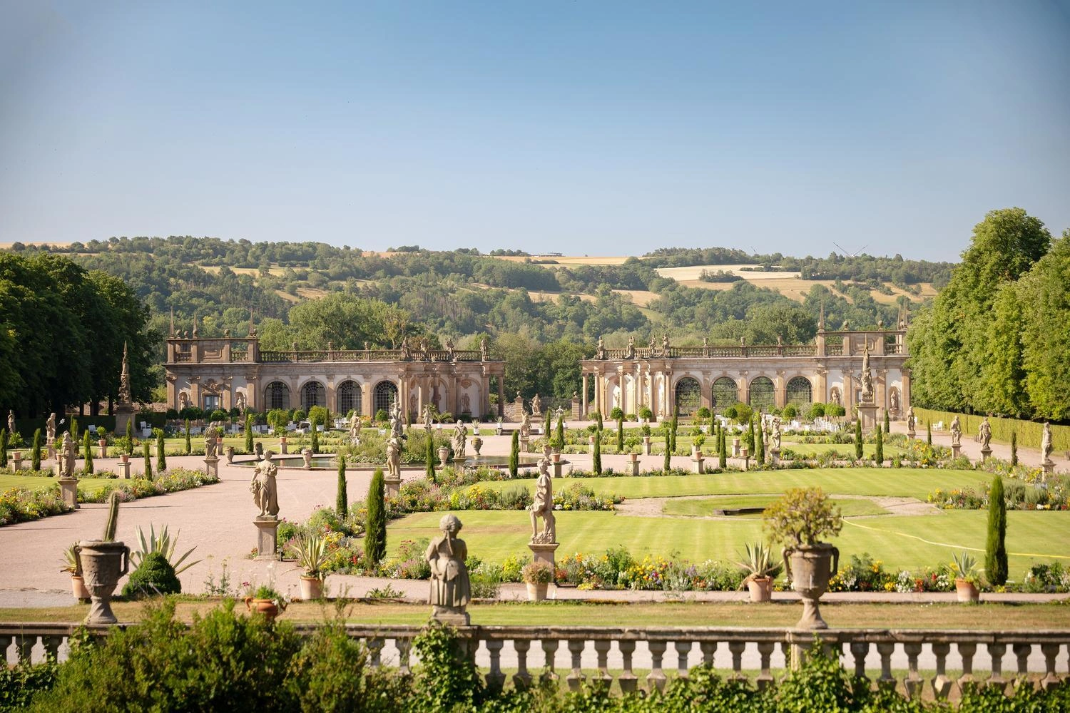 Barocker Schlossgarten mit symmetrischen Blumenbeeten, Skulpturen und Orangerie im Hintergrund, umgeben von grünen Hügeln unter blauem Himmel.