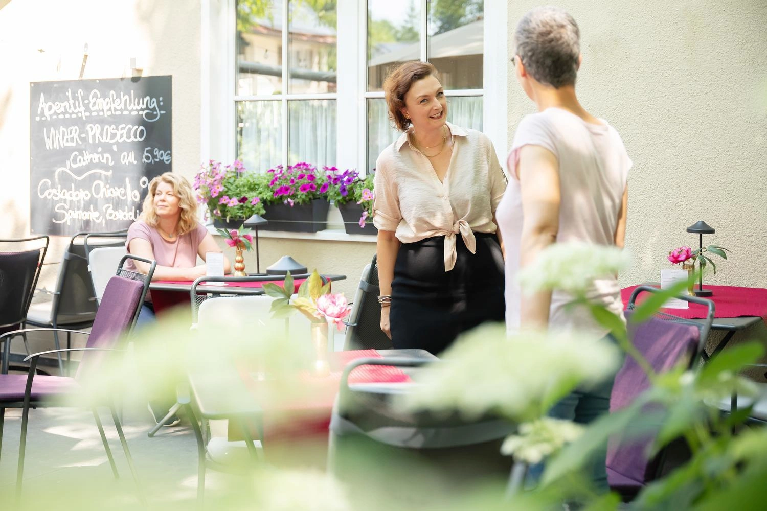 Gemütliche Caféterrasse mit Tischen, Blumen und einer Tafel mit Aperitif-Empfehlungen vor hellen Fenstern.