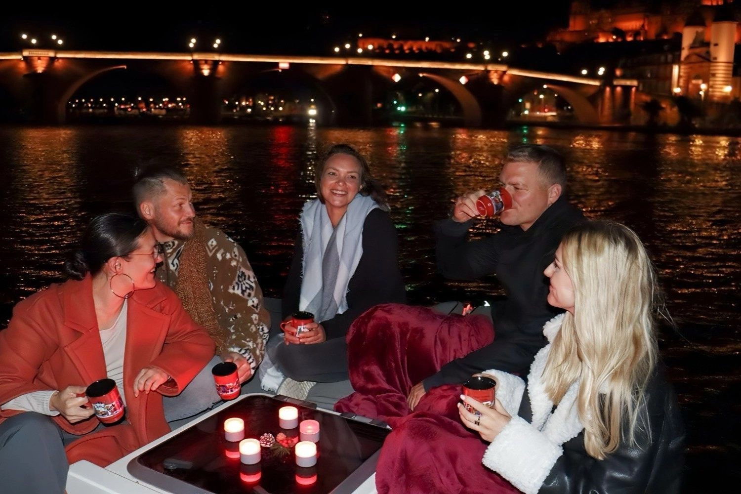 Gruppe sitzt nachts auf einem Boot, trinkt aus Tassen, umgeben von Kerzen, mit beleuchteter Brücke und Stadt im Hintergrund.