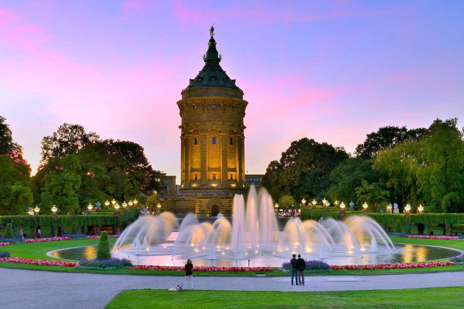 Ein Turm im lila-rosa Sonnenuntergang in einem Park. Davor ein Brunnen mit Wasserspielen.