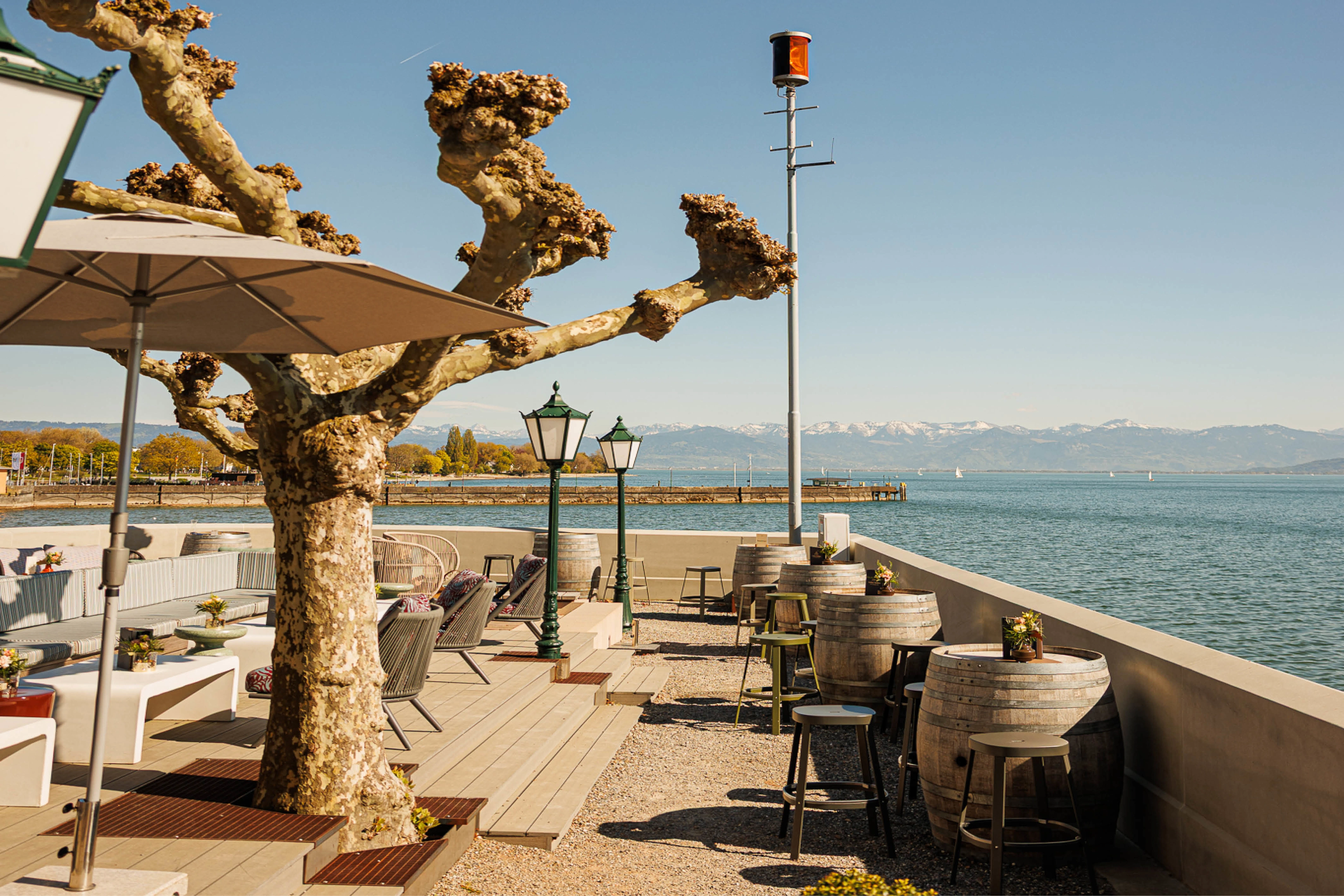 Terrasse mit Sitzplätzen und Weinfässern direkt am See, Blick auf Berge und Steg; steht für Gastronomie mit Seeblick und entspannte Auszeit am Wasser.