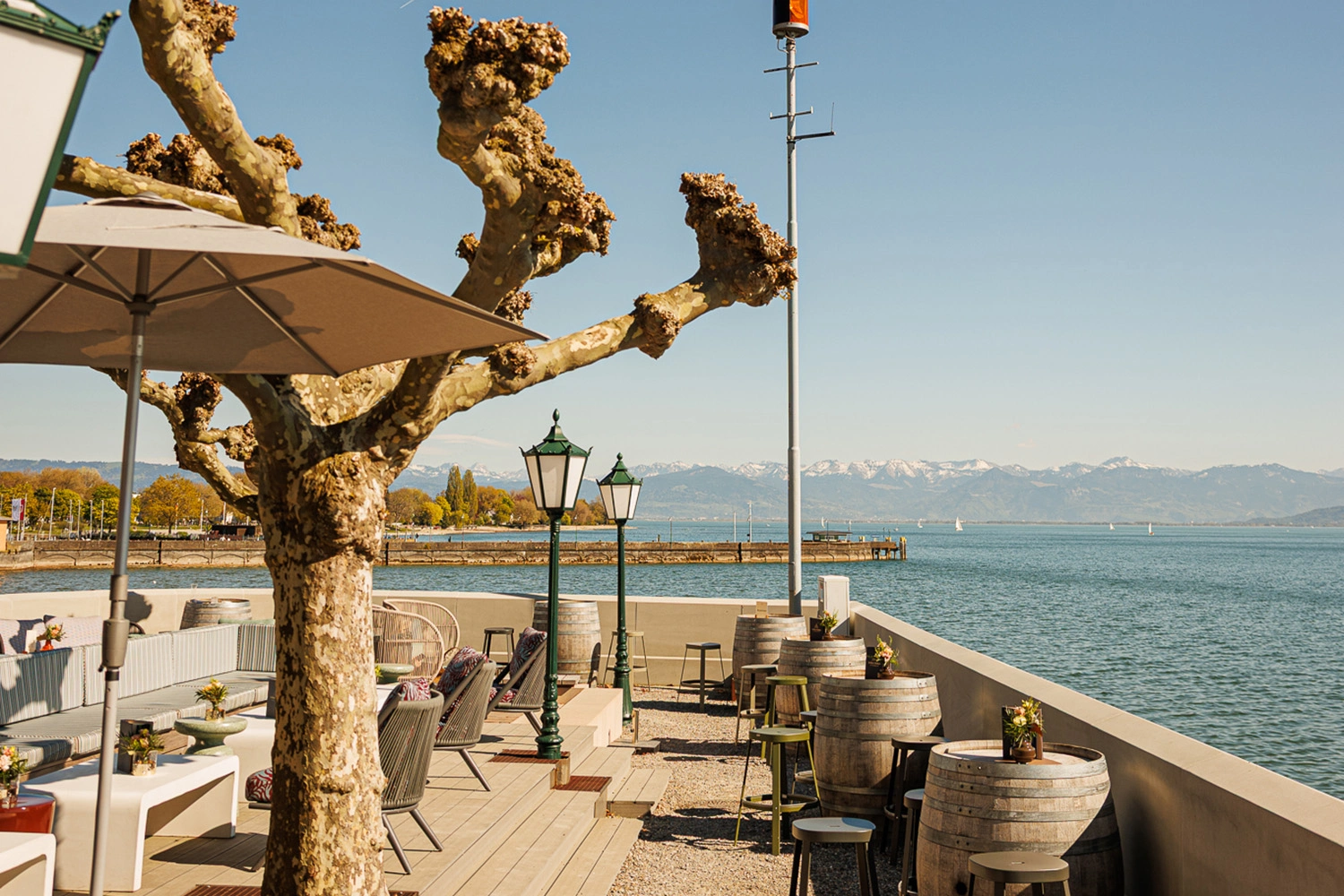 Terrasse mit Sitzplätzen und Weinfässern direkt am See, Blick auf Berge und Steg; steht für Gastronomie mit Seeblick und entspannte Auszeit am Wasser.