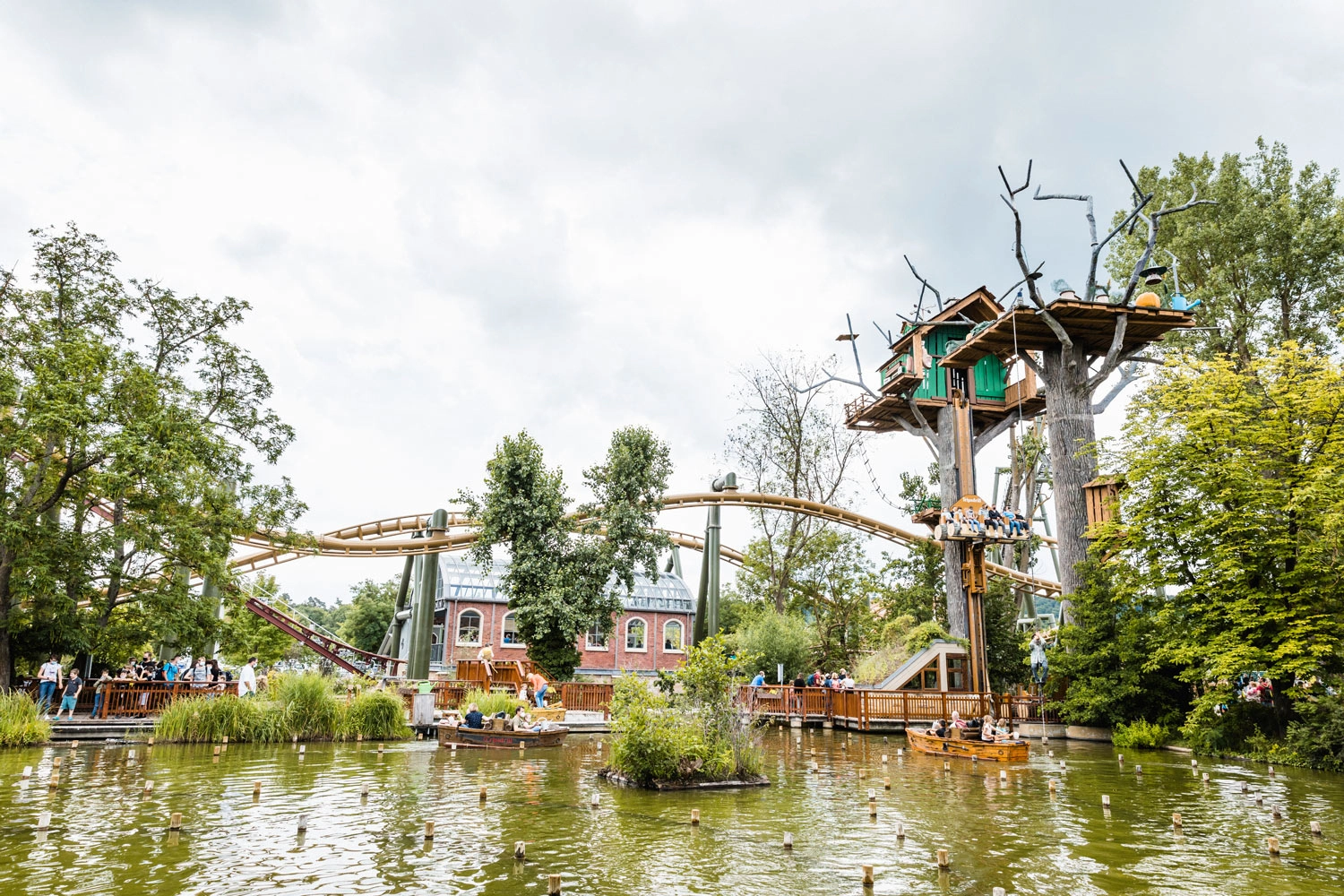 Blick auf den kleinen See in einem Freizeitpark, daneben Baumhäuser, im Hintergrund Achterbahnen.