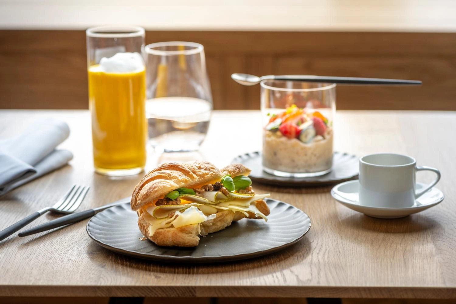 Frühstück mit Croissant-Sandwich, frischem Orangensaft, Kaffee und Overnight-Oats mit Früchten auf einem gedeckten Holztisch.