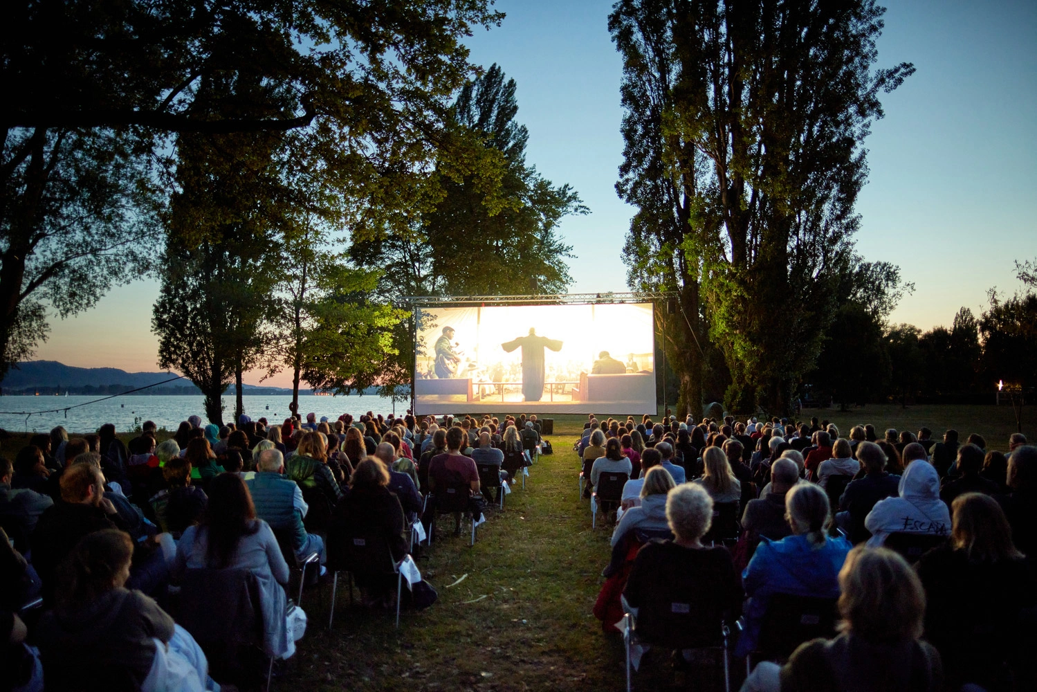 A large cinema screen stands on the lakeshore. People sit in two rows in front of it and watch a film together as dusk falls.