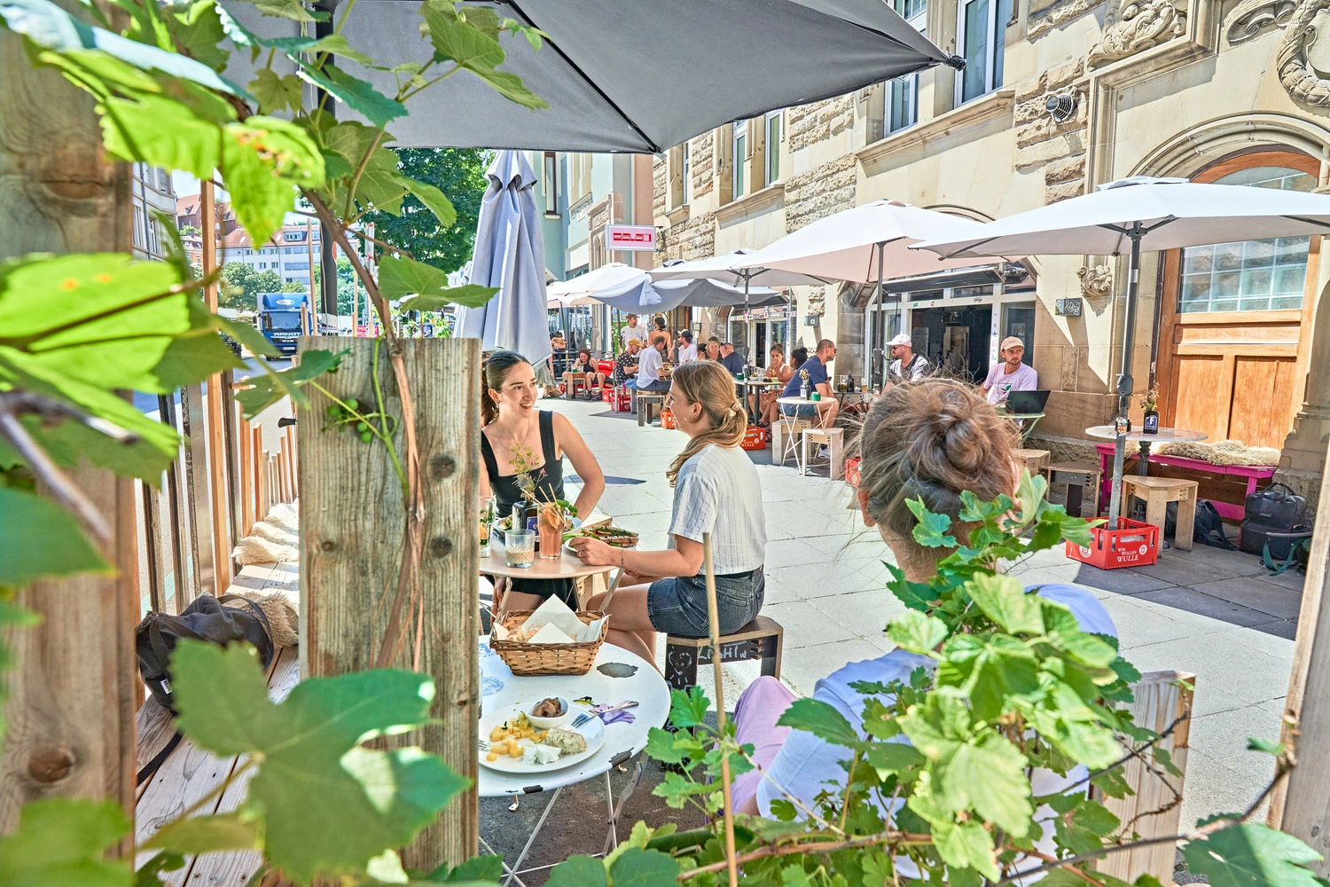 Straßencafé mit Sonnenschirmen und Gästen, die an Tischen essen und trinken, umgeben von Pflanzen, vor historischen Gebäuden in Stuttgart.
