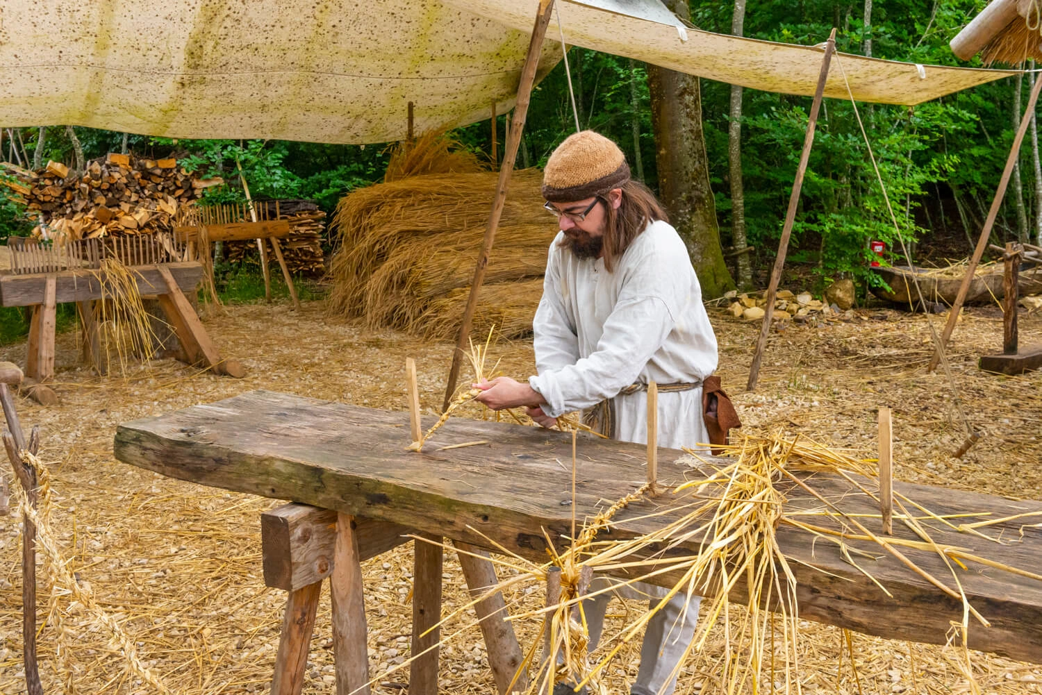 A man in medieval clothing stands in front of a wooden rack and twists straw.