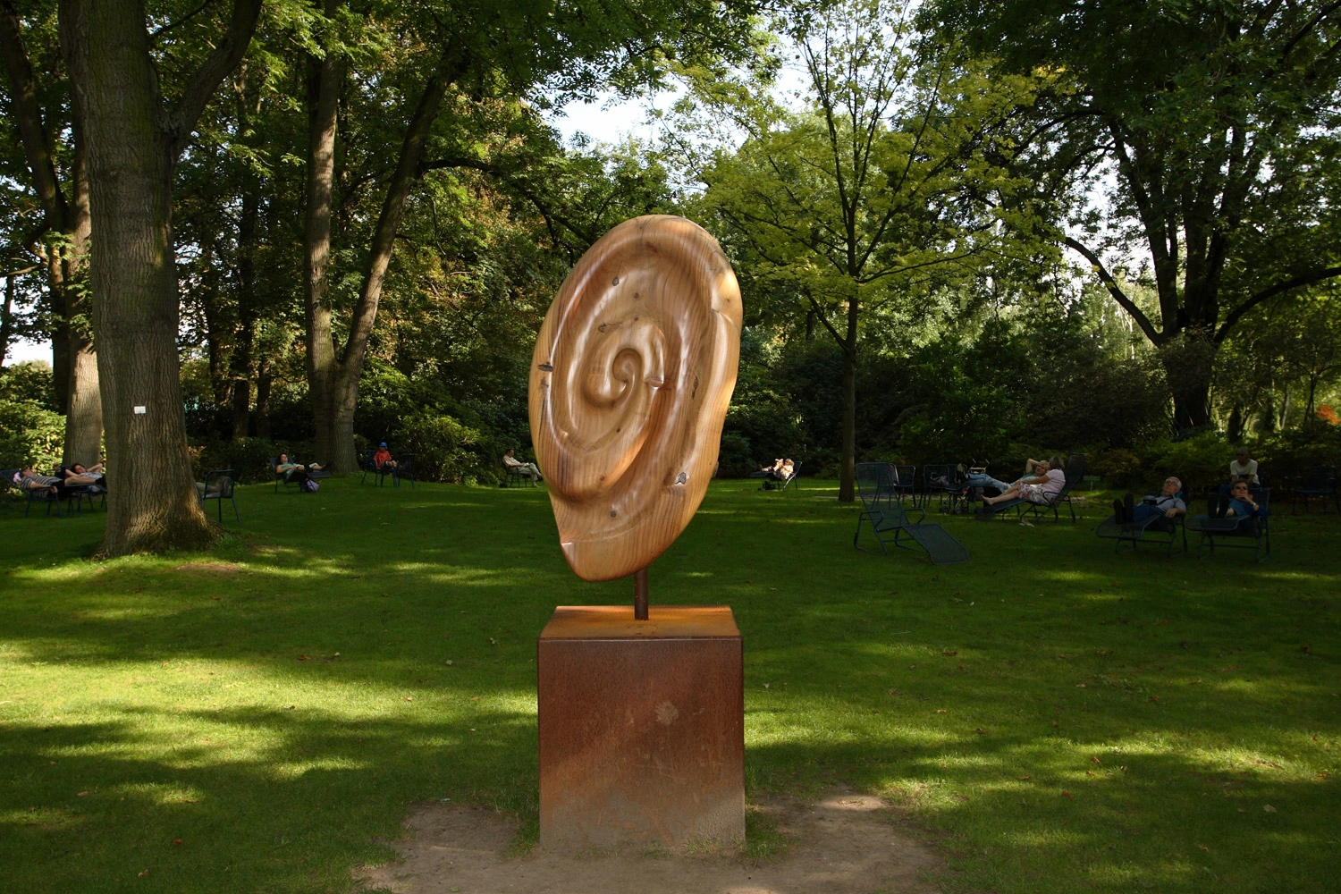 A wooden sculpture in the shape of an ear stands in a green park, surrounded by trees and relaxed visitors.