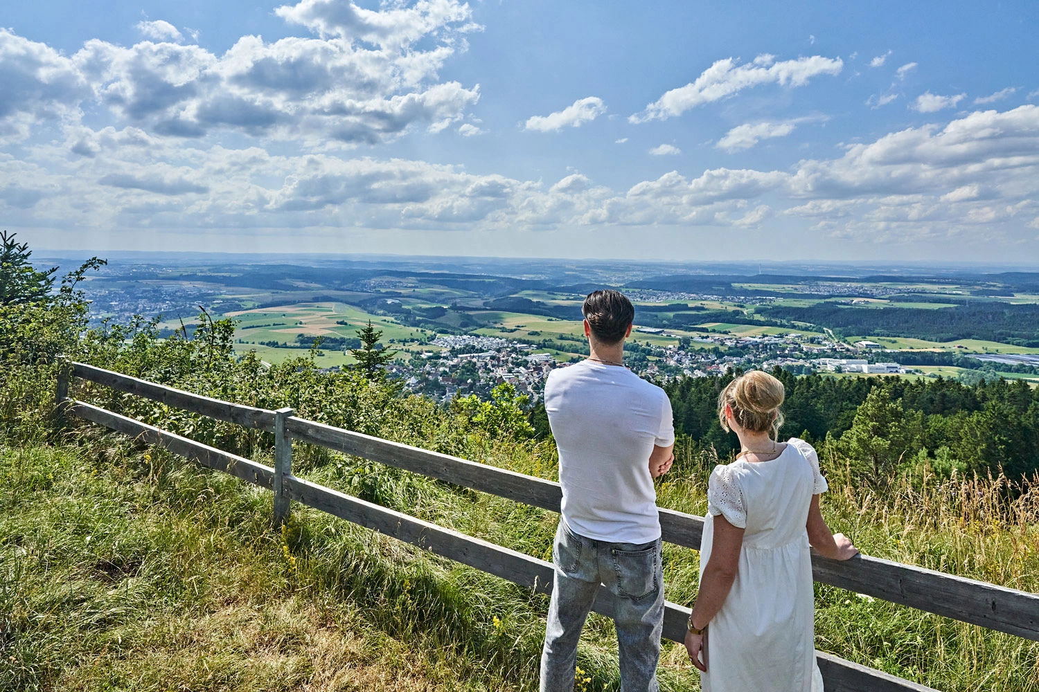 Zwei Personen stehen an einem Holzgeländer und blicken über Felder, Wälder und Dörfer der Schwäbischen Alb unter blauem Himmel mit Wolken.