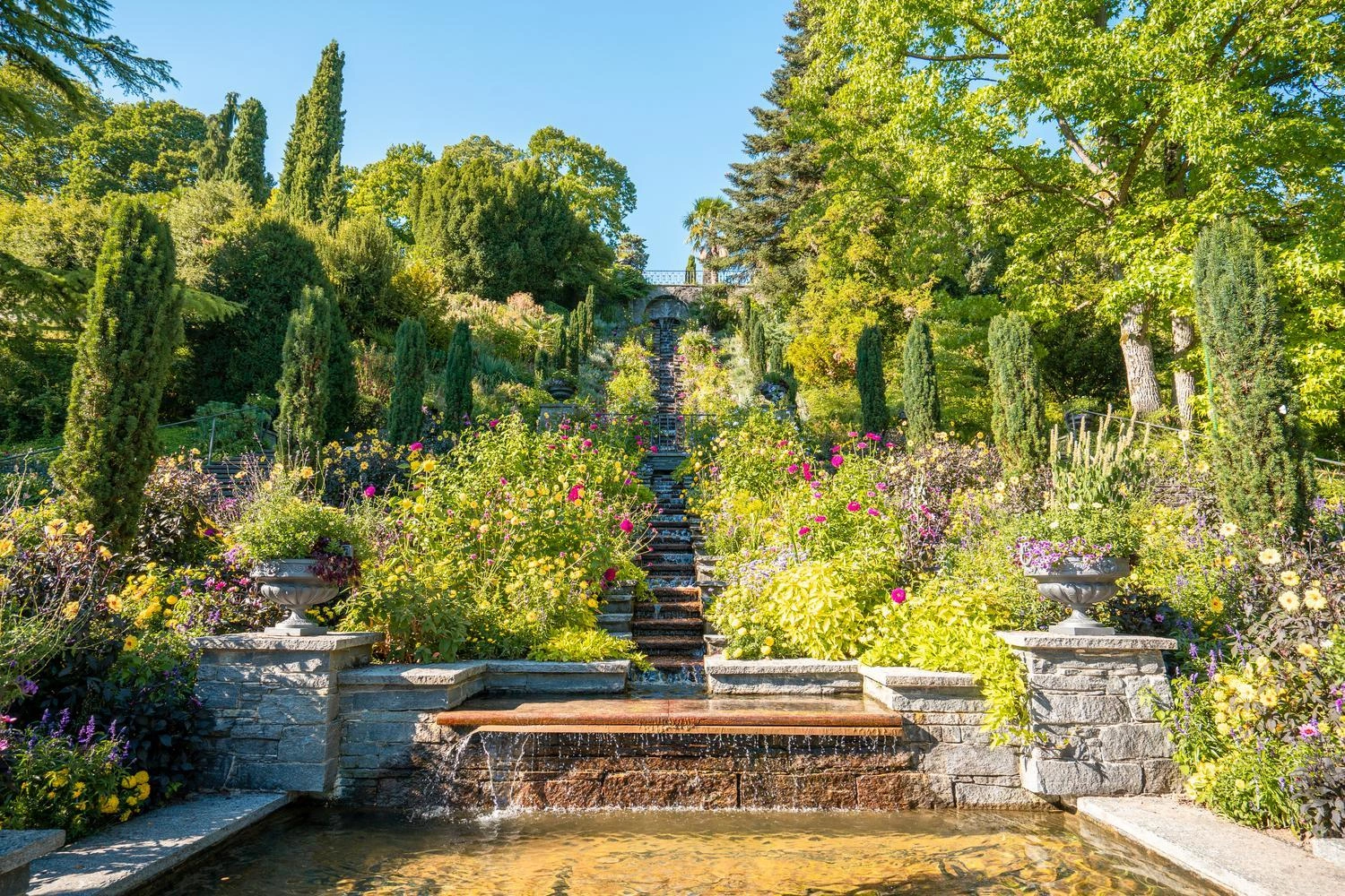 Steinerne Wassertreppe umgeben von bunten Blumen und hohen Bäumen, die in einen terrassierten Garten auf der Insel Mainau führt.
