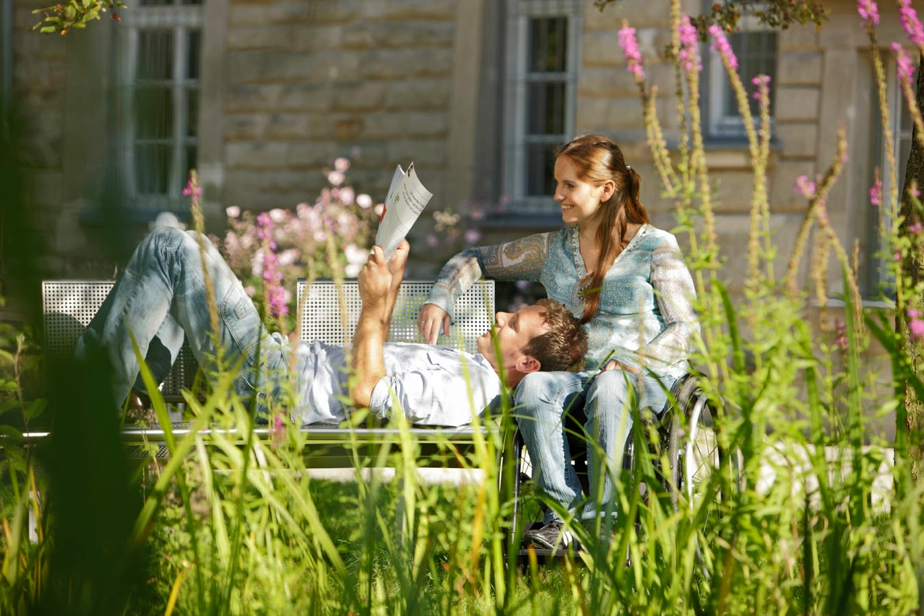 A man is lying on a park bench. A woman in a wheelchair is sitting next to him. He has rested his head on her lap and is reading the newspaper.