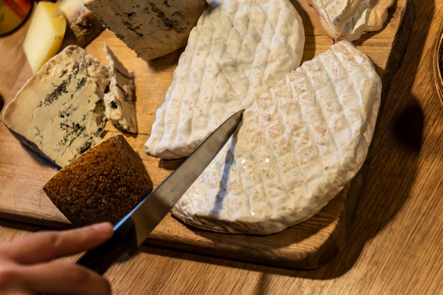 There are different kinds of cheese on a wooden board. A person cuts off a piece of cheese with a knife.