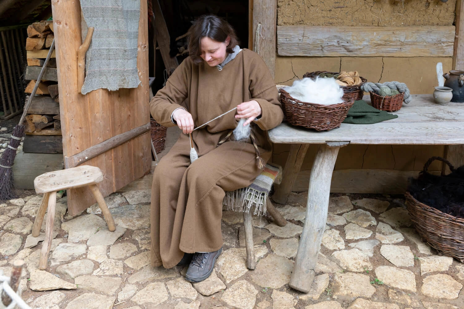 A woman in medieval clothing sits at a table in front of a hut, preparing yarn for weaving.