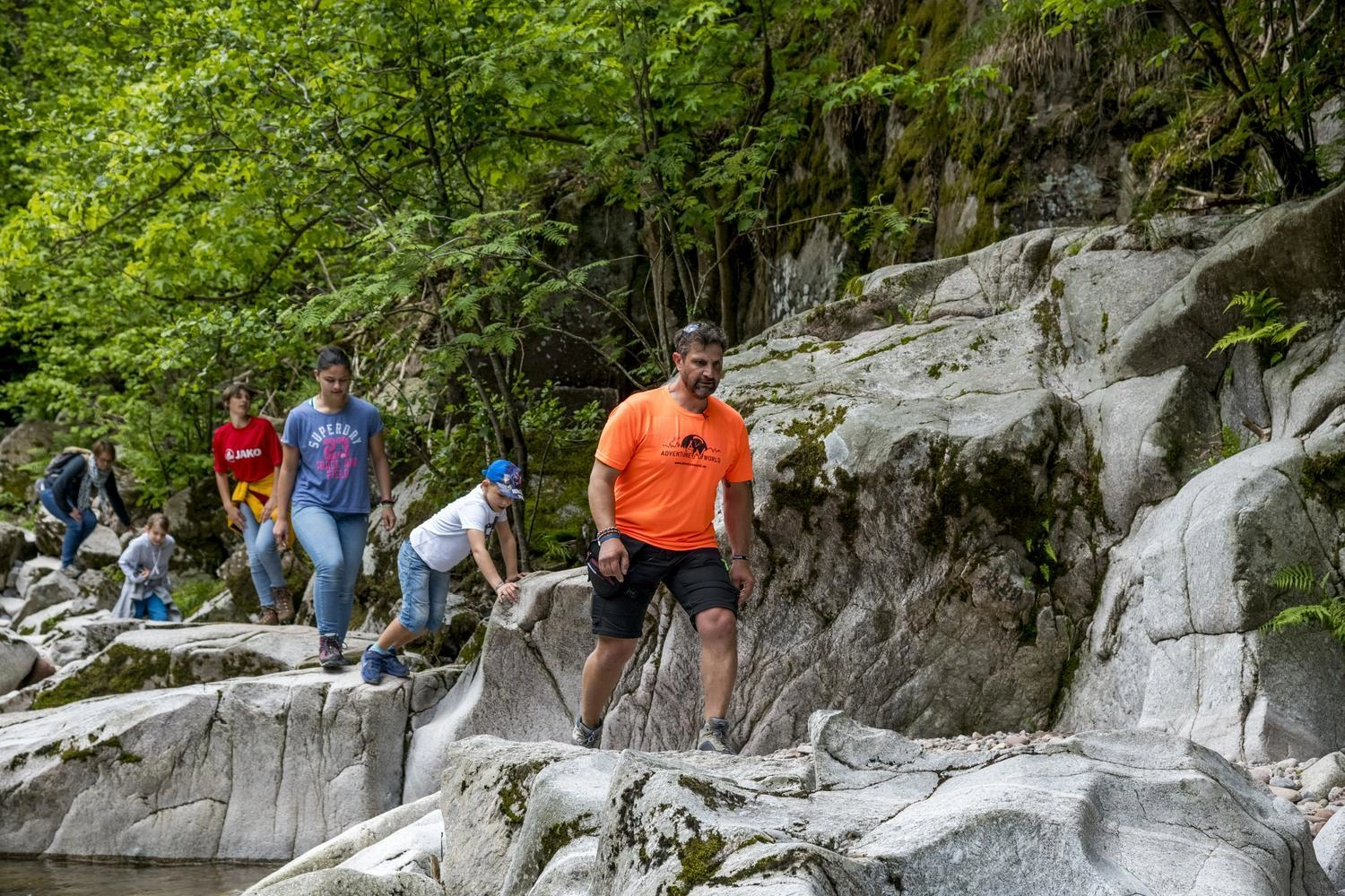 A guide leads a group of children and adults over large stones and rocks along a river.